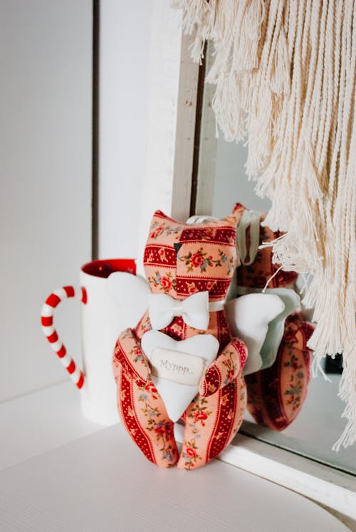 Soft cat shaped toy with bow tie and heart in hands placed in table near mug with candy cane handle