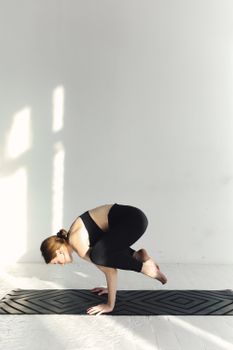 Woman in black activewear practicing crow pose on a yoga mat indoors, emphasizing balance and mindfulness.
