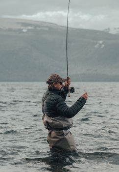 An adult fly fisherman standing knee-deep in a mountain lake, casting a fishing line.