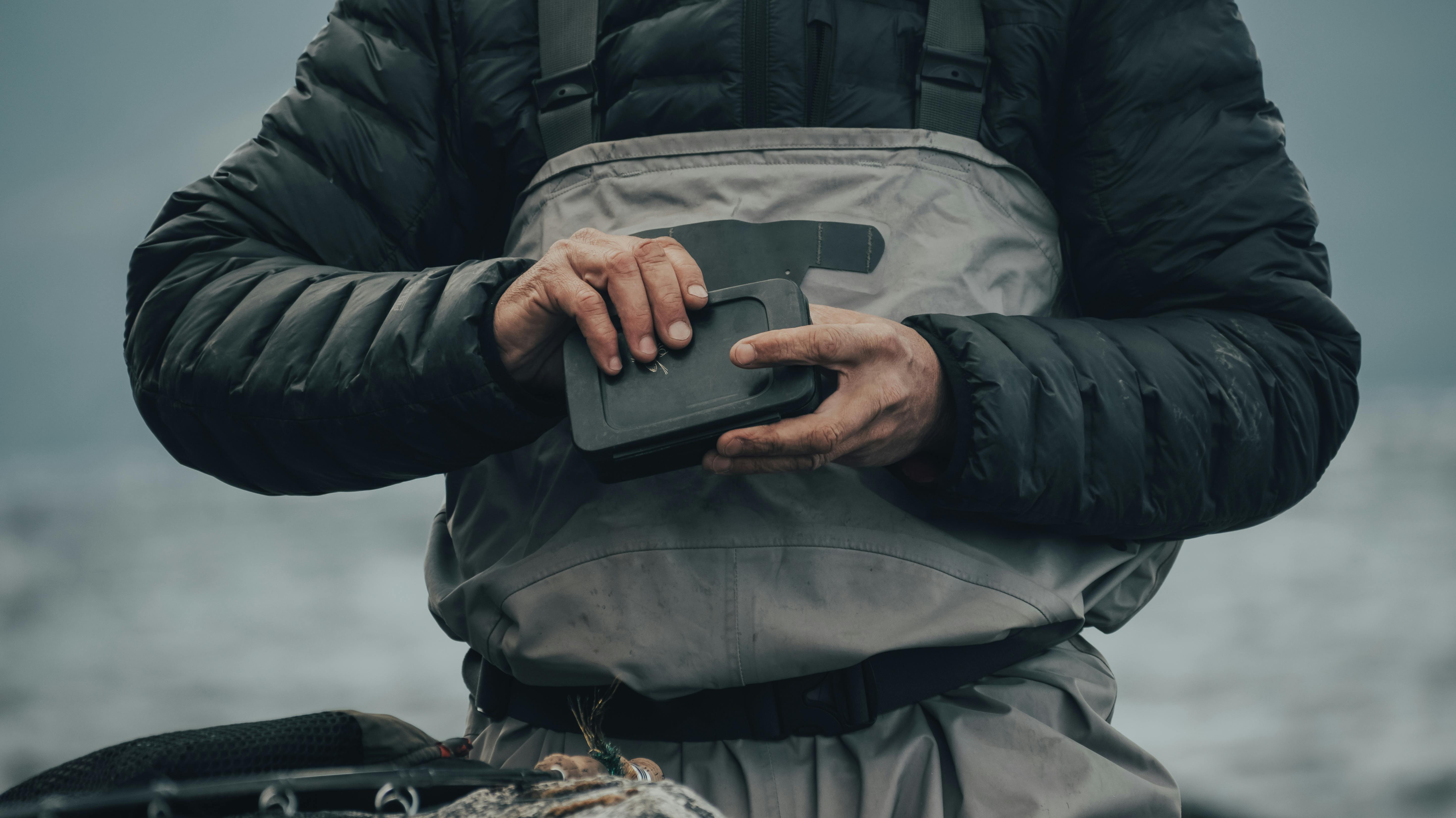A detailed view of hands holding a fly fishing reel box outdoors by the water.