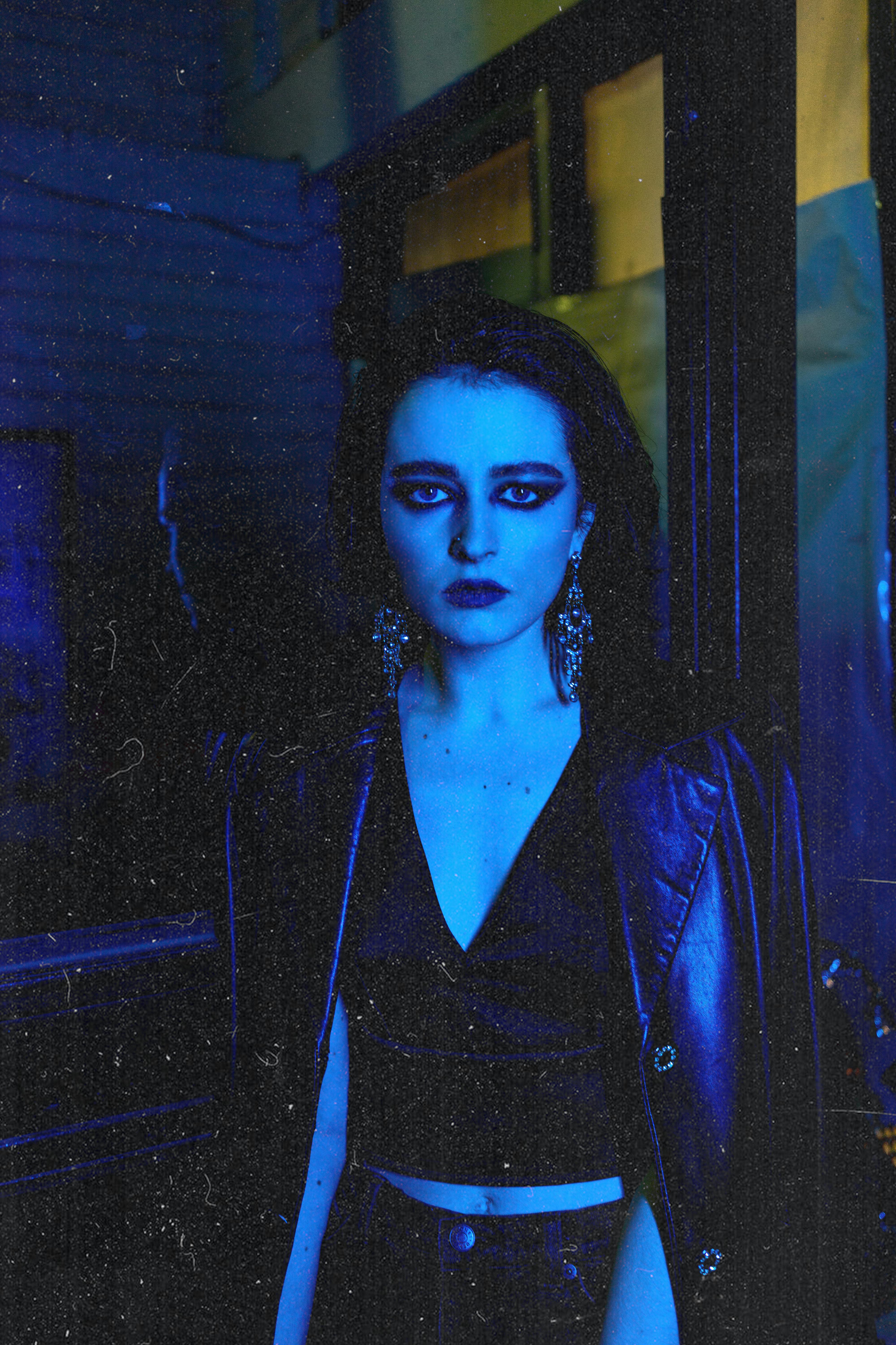 Slender woman in elegant leather jacket and earrings looking at camera in blue light on dark street