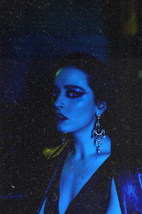 Eccentric female in sparkling earring looking at camera in darkness with blue neon light