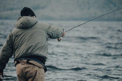 Free Fisherman casting with a fishing rod wearing a gray jacket by the ocean. Stock Photo