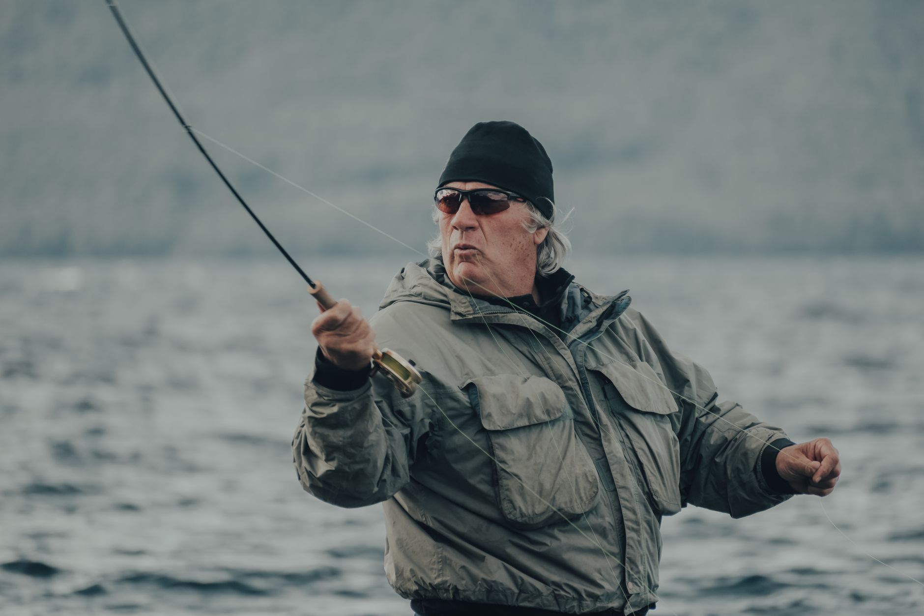 Elderly man fishing by the lakeside, wearing warm attire and sunglasses, casting his line.