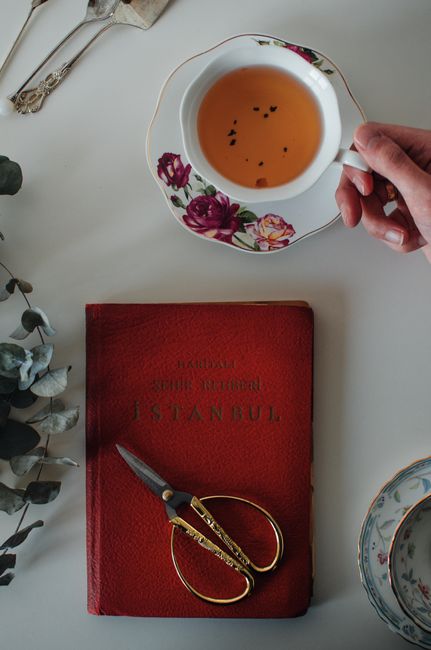 Overhead view of crop faceless woman at table with tea cup with saucer near scissors on red book near plant with green leaves near cutlery in bright room
