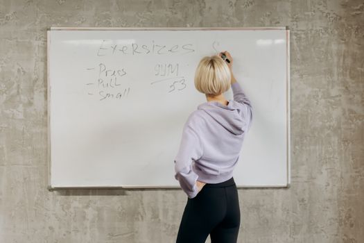 Female writing workout schedule on whiteboard in gym setting.