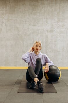A focused woman in sportswear sitting with a medicine ball in a gym setting.