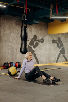 Blonde woman sitting on a workout mat in a modern gym surrounded by sports equipment.