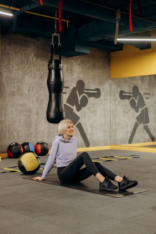 Free Blonde woman sitting on a workout mat in a modern gym surrounded by sports equipment. Stock Photo
