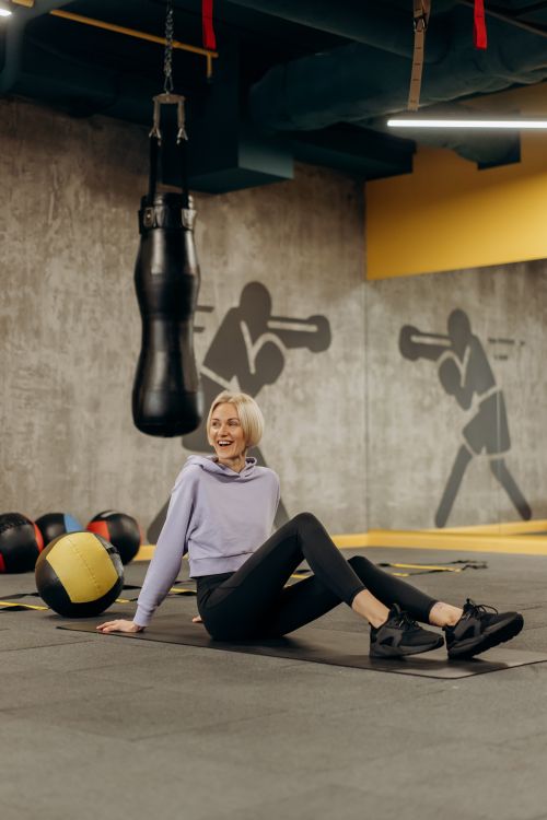 Free A woman relaxes on a gym floor surrounded by boxing and fitness equipment, smiling warmly. Stock Photo