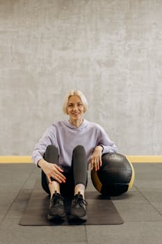 Woman sitting on mat in gym, resting with medicine ball, promoting active lifestyle and fitness.