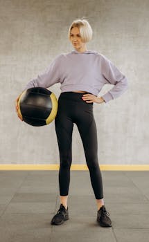 A fit young woman wearing athleisure holding a medicine ball in a fitness studio.