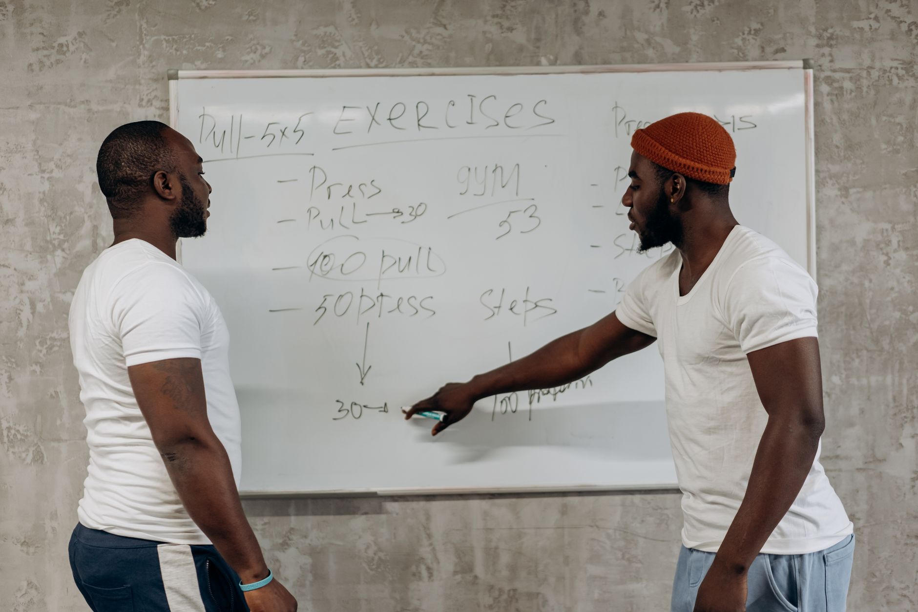 健美訓練提升自信:從新手到自我肯定的實用步驟 Two men discussing exercise routines on a whiteboard, planning gym workouts indoors.