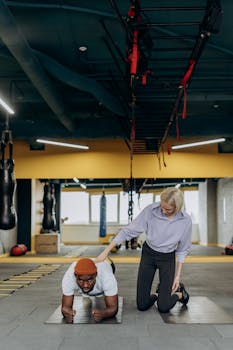 A woman coach guides a man doing a plank exercise in an indoor gym setting.