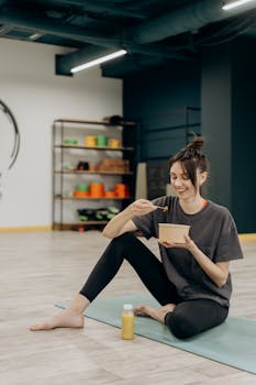 A smiling woman sitting on a mat enjoying a healthy meal indoors after exercise.