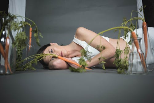 Woman in White Tank Top Lying on Floor with Carrot on her Arm