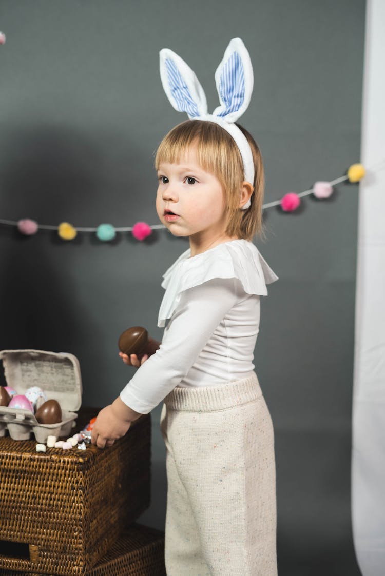 A Girl Wearing Blue And White Bunny Ears Headband Standing While Holding A Chocolate Egg