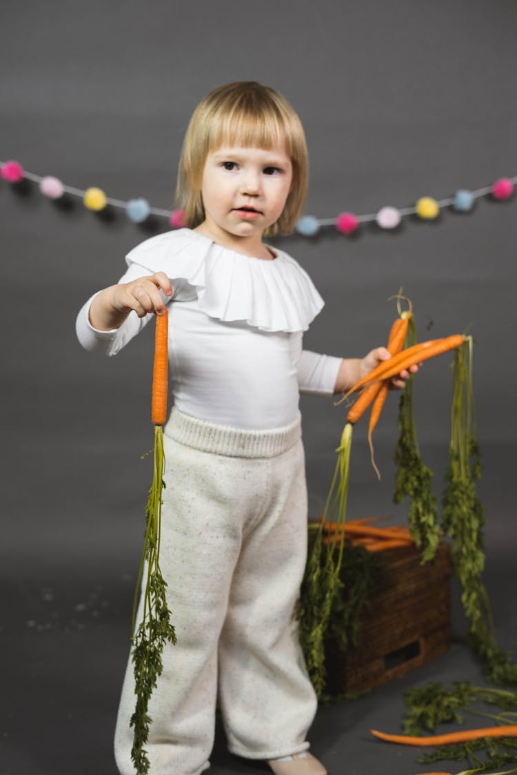 A Kid Holding Carrots While Standing Near A Gray Background