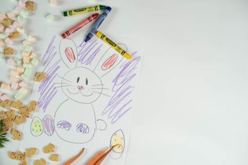 Top view of colorful crayons and picture of bunny and eggs placed on white background with marshmallows and cookies during Easter holiday