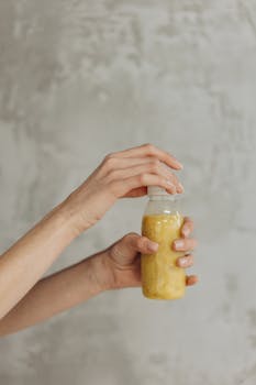 Close-up of a woman's hand holding a refreshing yellow juice bottle against a neutral background.
