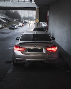 A silver BMW 4 Series coupe parked under an overpass on a wet, rainy street during dusk.