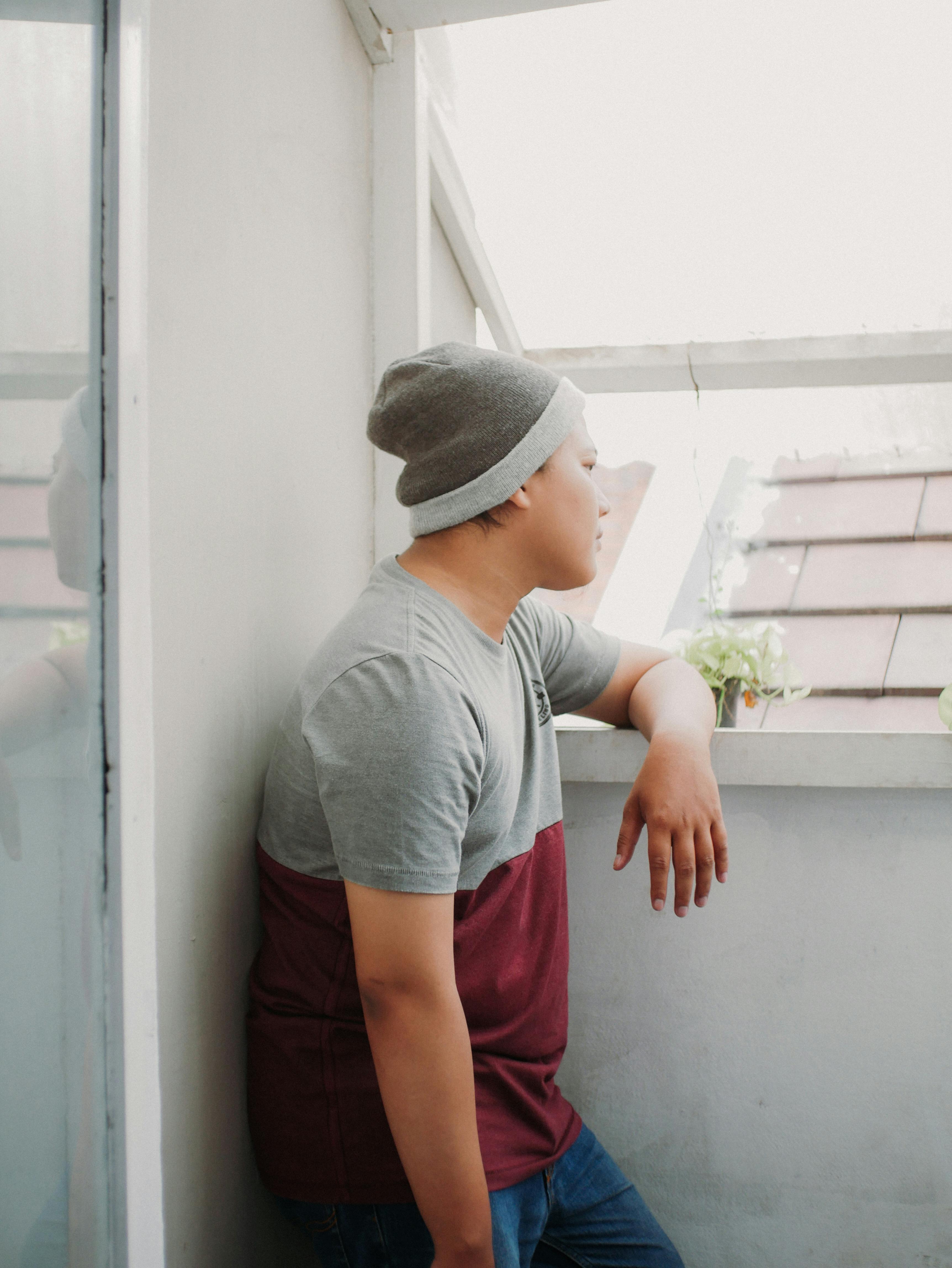 Free A man in casual attire with a beanie, standing indoors by a window, deep in thought. Stock Photo