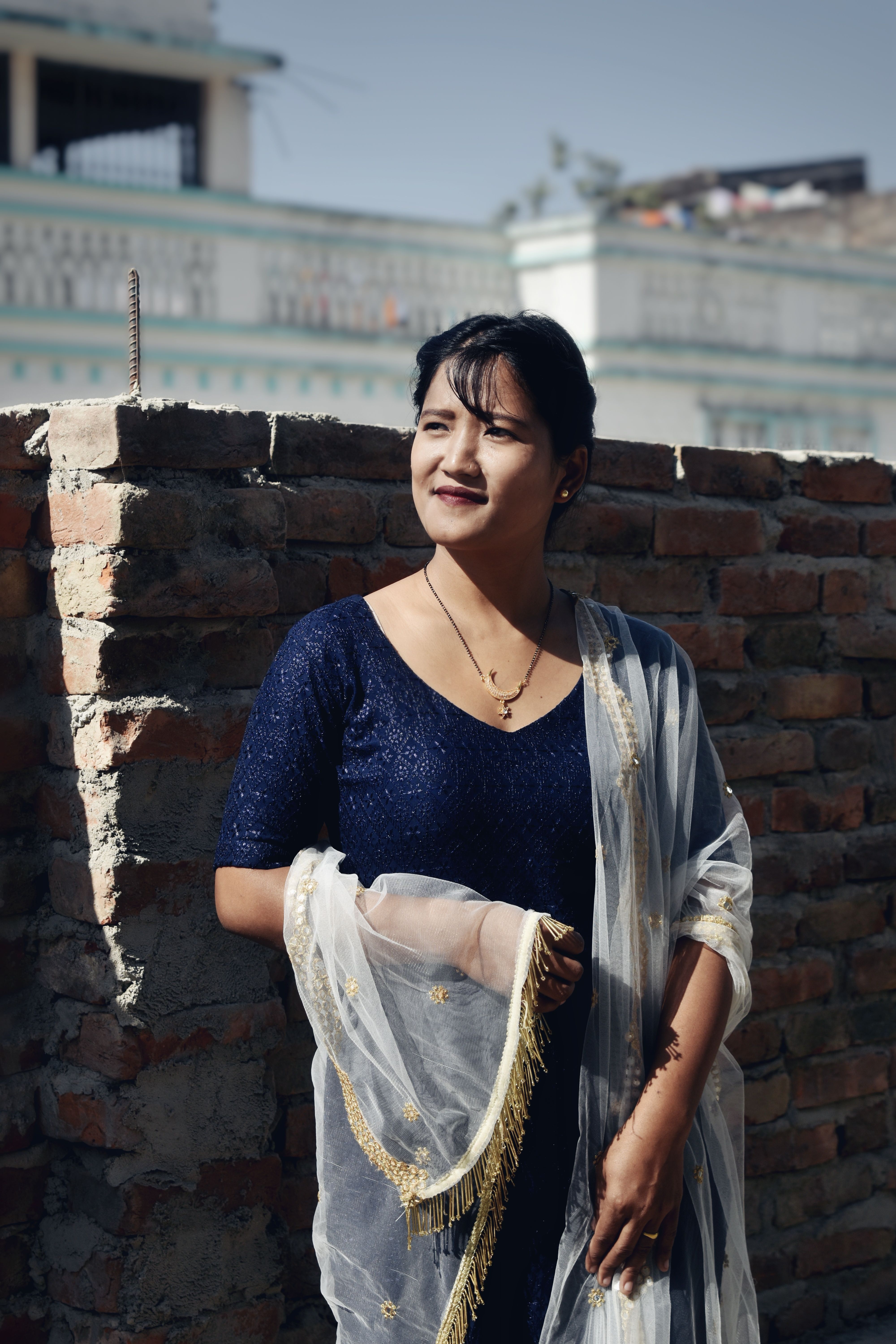 A woman in a blue dress smiles while standing outdoors by a brick wall.