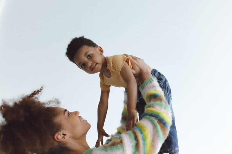 A Black mother lifts her smiling toddler son in a bright room, a tender moment between a parent and a young child.