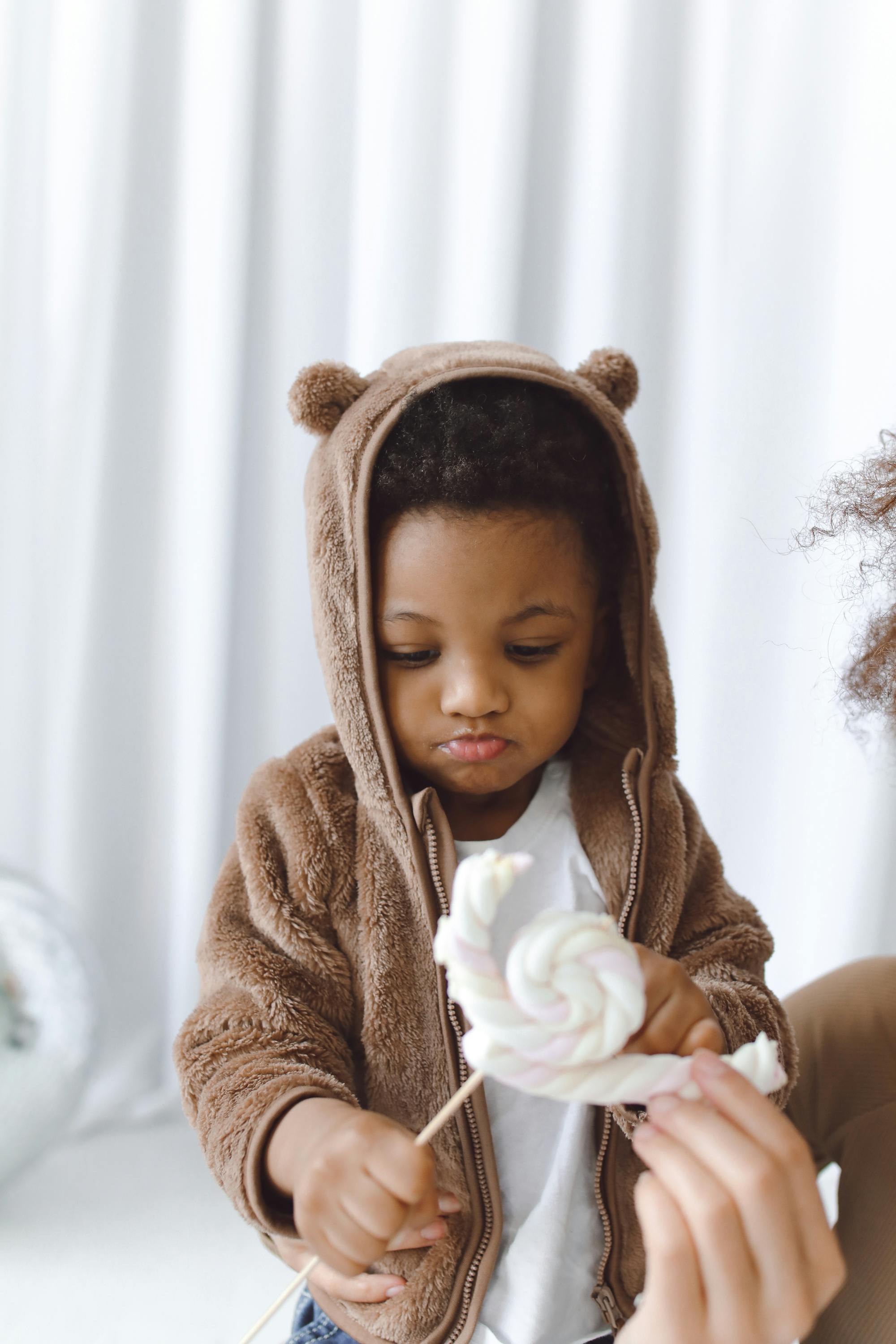 boy in brown hoodie jacket looking at a marshmallow candy on stick