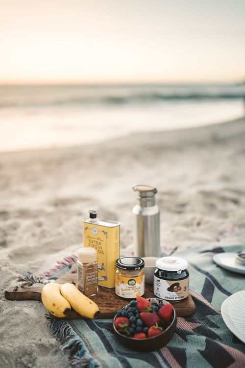 Free A beach picnic setup with fruits and essentials on a mat at sunset. Ideal for relaxing and enjoying the sea breeze. Stock Photo