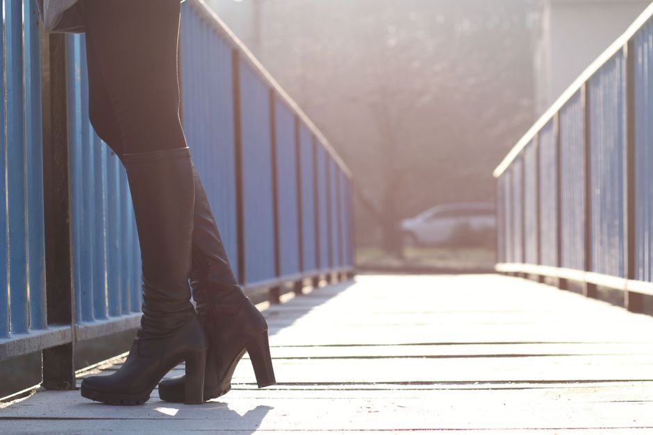 Close-up of black knee-high boots with block heels worn on a pedestrian bridge, showing sleek footwear that modernizes a tweed mini skirt