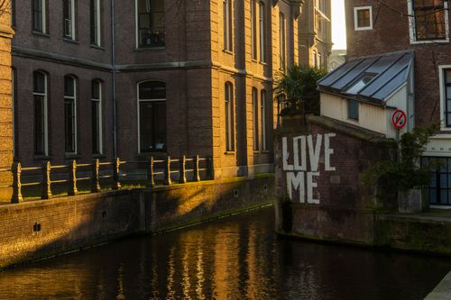 Free Scenic view of urban canal with 'Love Me' graffiti and sunlight casting warm hues on historic buildings. Stock Photo