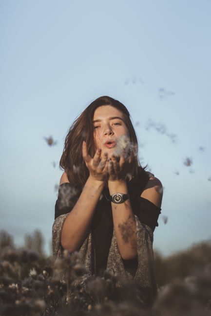 Happy young female with long dark hair in casual clothes standing in meadow and blowing dandelion towards camera