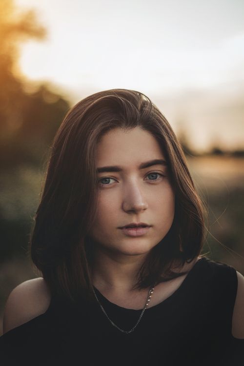 Free Self assured young lady with dark hair in casual clothes looking at camera pensively while resting in countryside against sunset sky Stock Photo