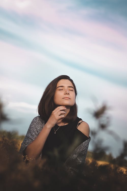 Free Low angle of feminine young woman with dark hair in casual clothes enjoying free time in rural meadow with closed eyes against cloudy sunset sky Stock Photo