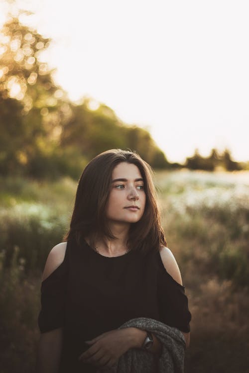 Dreamy young woman wearing black blouse while standing near field with plants and grass near trees and looking away in nature in daylight