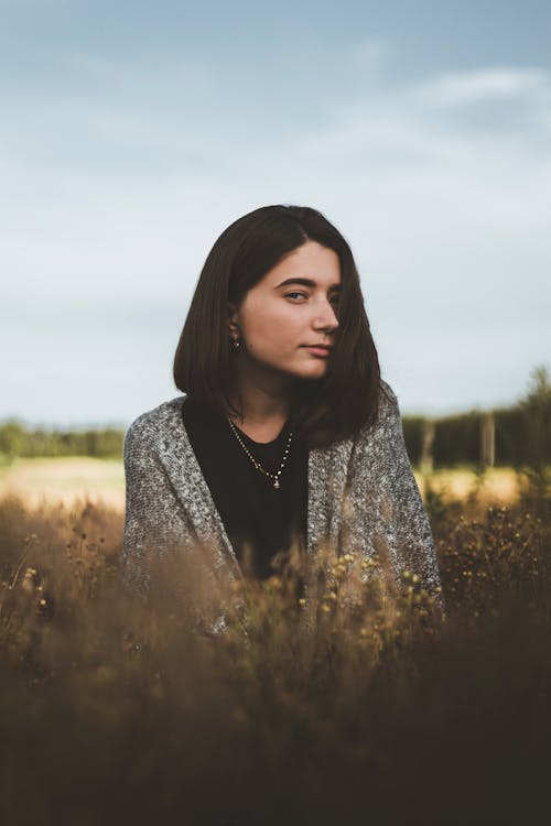 Young female with dark hair in casual clothes looking at camera while standing in meadow in countryside in autumn