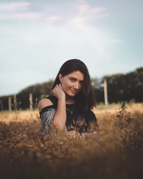 Young dreamy female with long dark hair bare shoulder in casual clothes touching neck and looking at camera while standing in dry grass in countryside in autumn