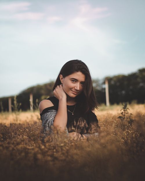 Free Young dreamy female with long dark hair bare shoulder in casual clothes touching neck and looking at camera while standing in dry grass in countryside in autumn Stock Photo