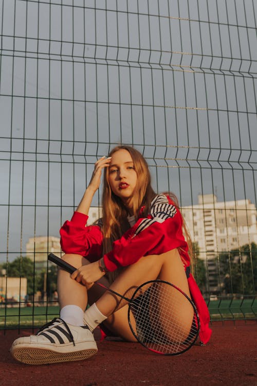Full body of young female with red lips and long hair in sportswear sitting on sports ground on tennis court near metal net railing and looking away while resting during badminton training