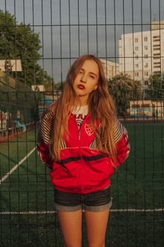 Young woman with long hair and red lips in stylish outfit standing on sports ground and looking at camera