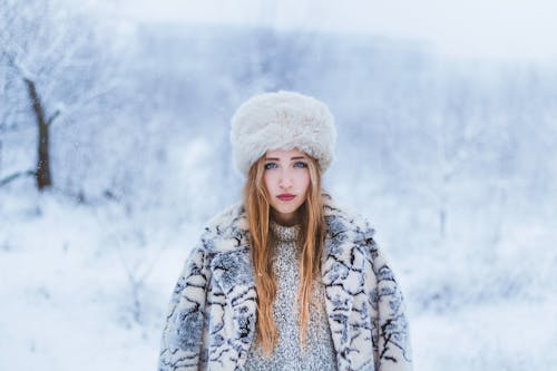 Young female with blue eyes in warm clothes and hat looking at camera while standing in snowy forest in winter