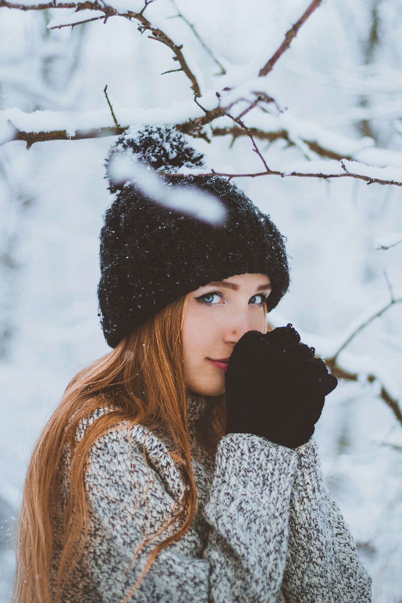 Woman in a snowy forest wearing a black beanie, mittens, and a gray marled knit sweater