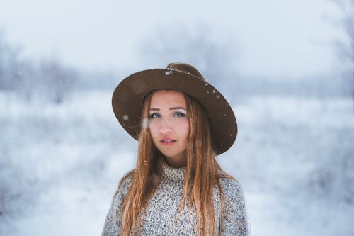 Young female in hat with wide brim and knitted sweater looking at camera while standing in snowy forest in countryside