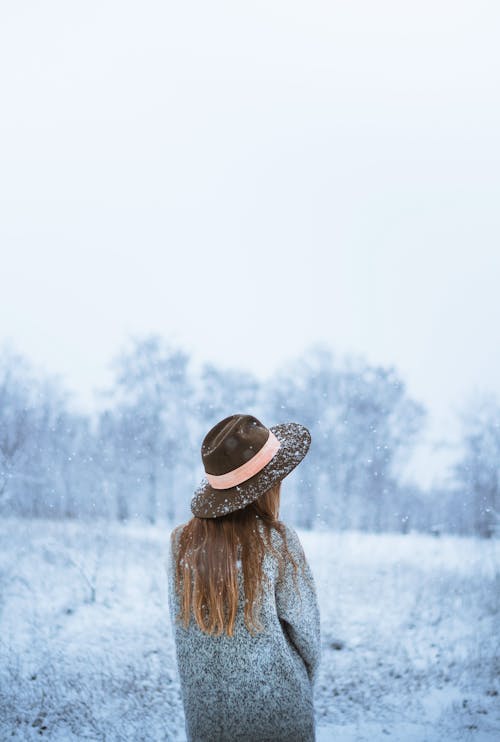 Back view of anonymous female in hat and outerwear standing in snowy valley in haze