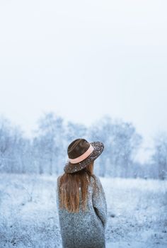 Back view of anonymous female in hat and outerwear standing in snowy valley in haze