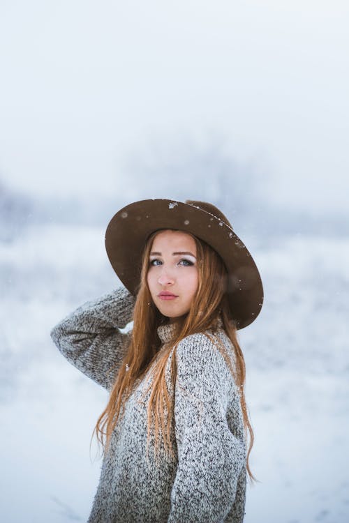 Side view of stylish young female wearing warm sweater with hand on hat standing against misty snowy terrain