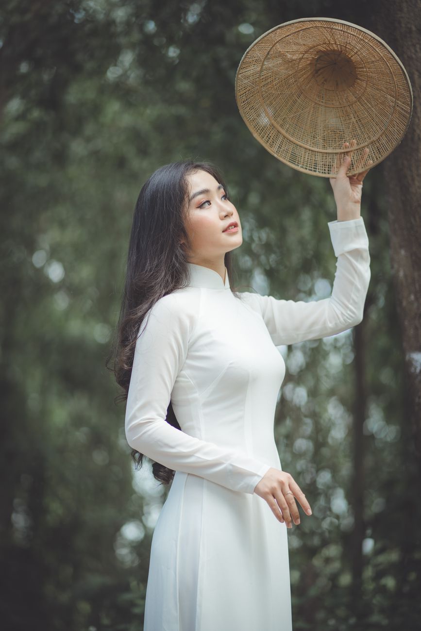 Elegant woman in a long white dress, holding a woven hat amidst lush greenery, looking serene.