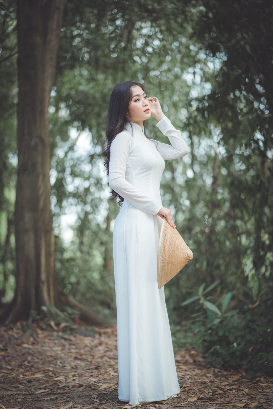 Asian woman in white traditional dress with a hat in a serene forest setting.