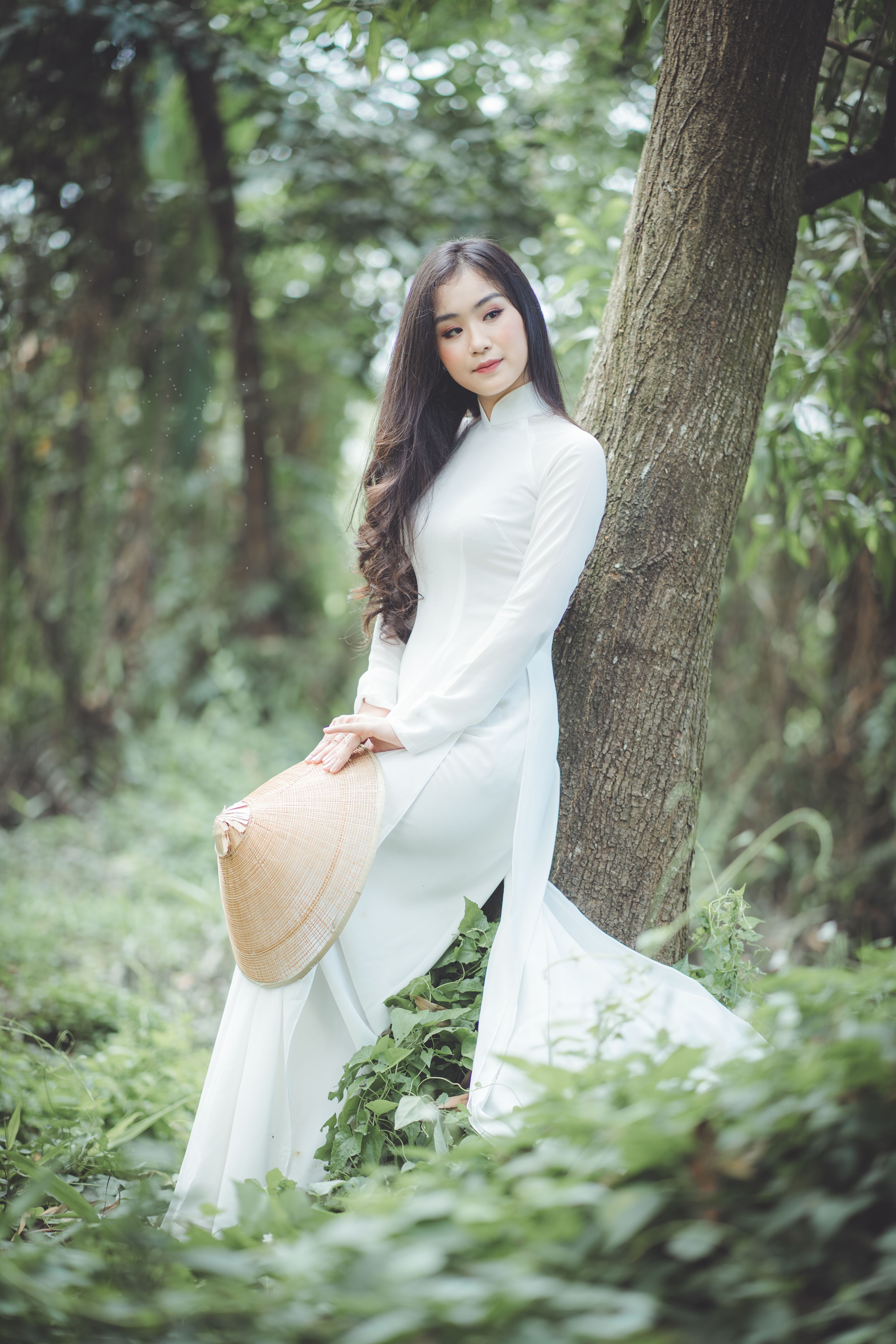 Asian woman in traditional dress leaning against a tree, holding a hat in a natural setting.